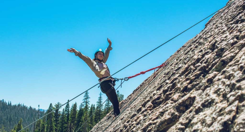 A person wearing a helmet is secured by ropes as they stand on a rocky incline and raise their arms in celebration. 